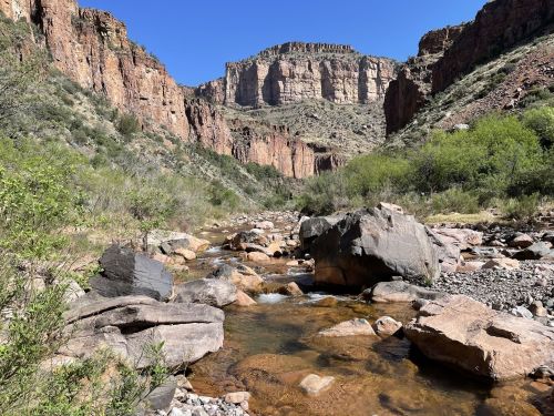 A rocky riverbed flows through a canyon with steep, rugged cliffs and green vegetation under a clear blue sky.