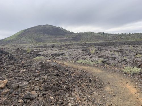 A rocky, volcanic landscape with a dirt path leading towards a distant hill under a cloudy sky.