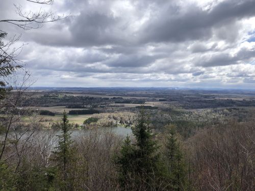 A scenic view from a height, showcasing a lake surrounded by trees and fields under a cloudy sky.
