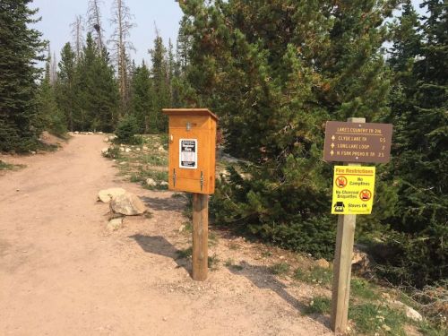 A dirt trail with a wooden information box and signs indicating nearby lakes and fire restrictions, surrounded by trees.