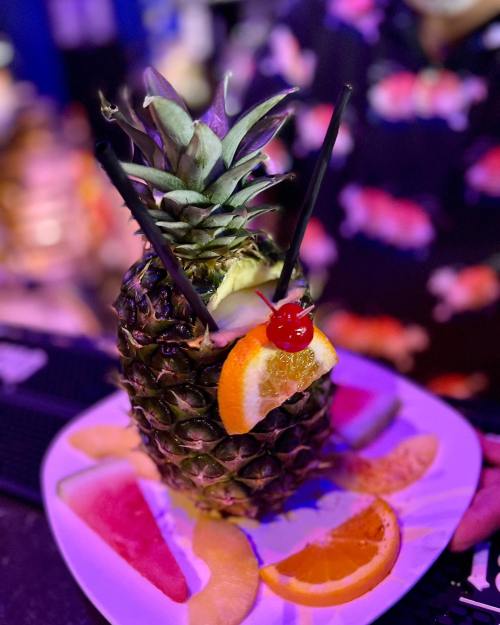 A pineapple cocktail garnished with fruit slices, straws, and a cherry, served on a plate.