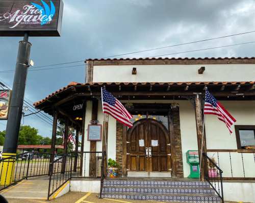 Exterior of a restaurant with a wooden door, American flags, and a sign that says "Open" under a cloudy sky.