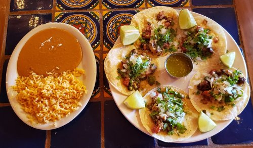 A plate of six tacos topped with cilantro and lime, served with rice and beans on a colorful tiled table.