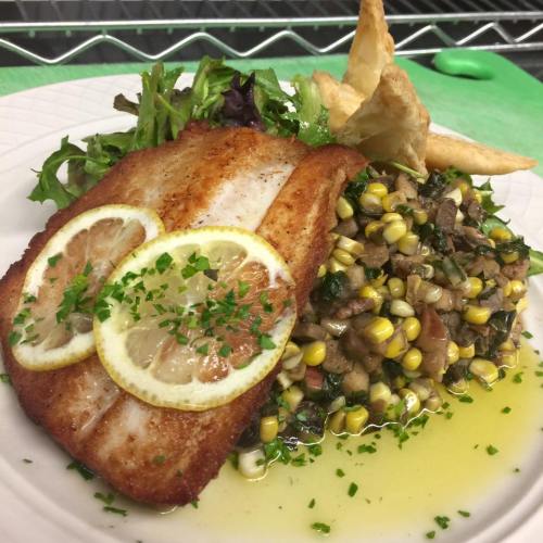 A plate featuring a fillet of fish with lemon slices, served with corn salad, greens, and crispy pastry triangles.