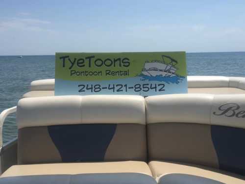 Sign on a pontoon boat advertising "TyeToons Pontoon Rental" with a phone number, set against a lake backdrop.