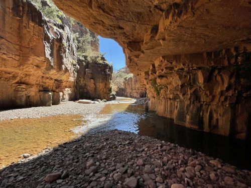 A rocky canyon with steep walls, a shallow river running through, and sunlight filtering in from above.