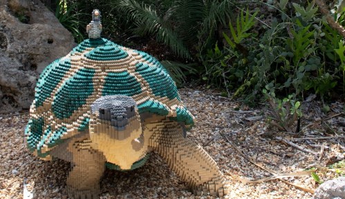 A large turtle sculpture made of colorful blocks, with a small figure on its back, set in a natural outdoor environment.