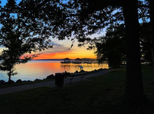A serene lakeside view at sunset, with a pier in the distance and silhouettes of trees framing the scene.