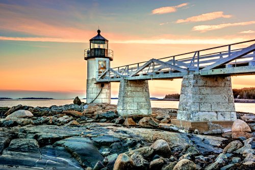 A lighthouse stands on a rocky shore at sunset, with a wooden walkway extending over the water.