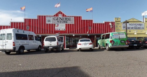 A colorful general store on Route 66, with various vehicles parked in front and flags waving above.