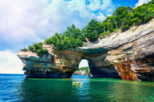 A scenic view of a natural rock arch over clear blue water, with kayakers exploring beneath it. Lush greenery surrounds the area.