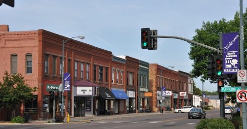 A quiet street scene featuring brick buildings, storefronts, and traffic lights in a small town.