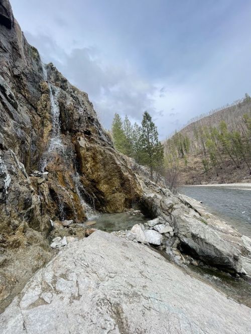 A rocky landscape featuring a waterfall cascading down a cliff, with a river and trees in the background under a cloudy sky.