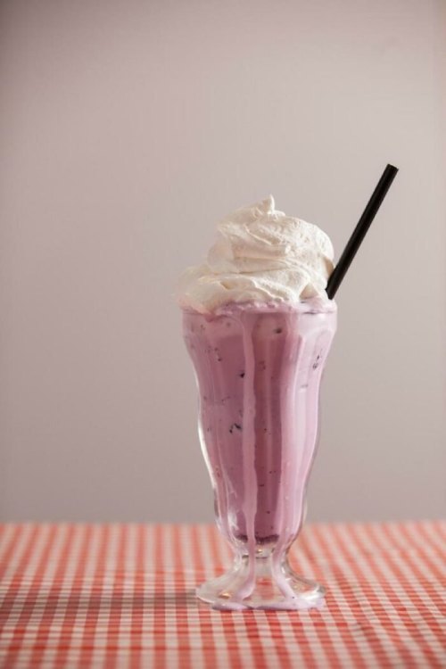 A tall glass of purple milkshake topped with whipped cream and a black straw, set on a checkered tablecloth.