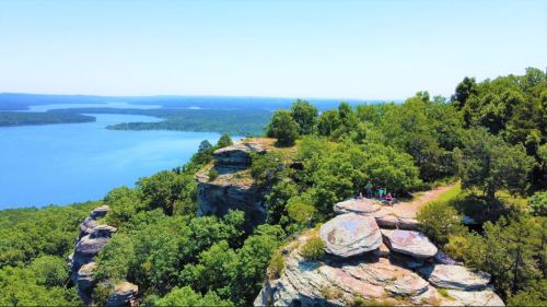 Aerial view of a rocky cliff overlooking a serene lake surrounded by lush greenery and clear blue skies.