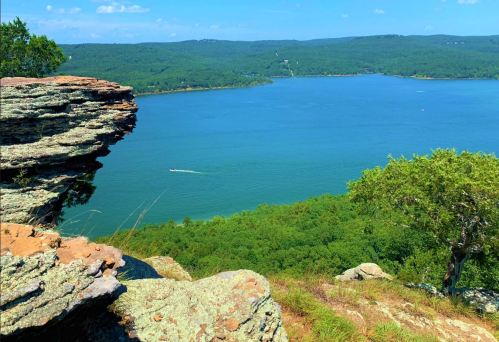 A scenic view of a lake surrounded by green hills, with rocky cliffs in the foreground and a clear blue sky.