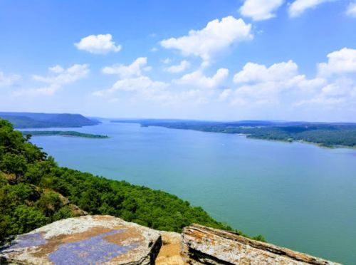 A scenic view of a wide river surrounded by lush green hills under a bright blue sky with fluffy clouds.