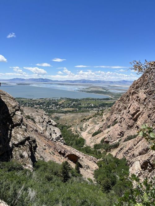 A scenic view of a valley with rocky cliffs, greenery, and a lake under a bright blue sky with scattered clouds.