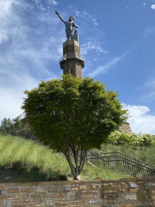 A tall statue atop a stone tower, surrounded by greenery and a blue sky with clouds.