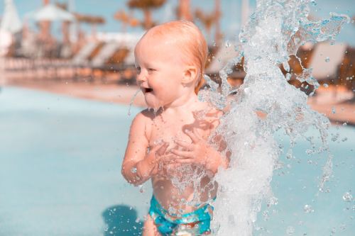 A joyful toddler splashes in water at a pool, surrounded by sunlight and playful water jets.