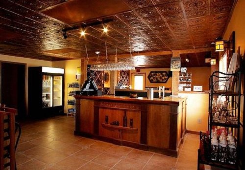 A cozy bar area with a wooden counter, shelves of drinks, and a decorative ceiling. Warm lighting enhances the ambiance.
