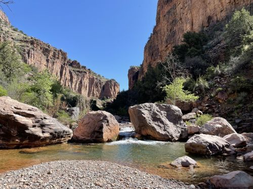 A serene river flows between rocky cliffs, surrounded by lush greenery under a clear blue sky.