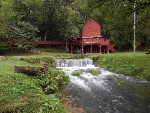 A red wooden mill beside a small waterfall, surrounded by lush greenery and trees.
