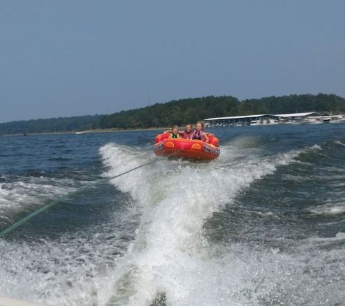 Three people enjoy tubing on a lake, being pulled by a boat, with waves splashing around them.