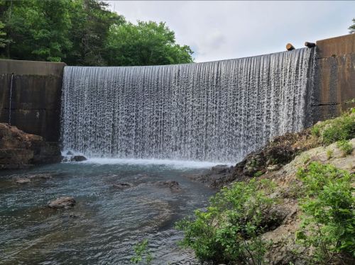 A waterfall cascading over a dam into a rocky river, surrounded by lush greenery and a cloudy sky.