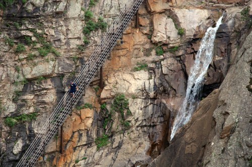 A person walks on a wooden bridge along a rocky cliff, with a waterfall cascading nearby.