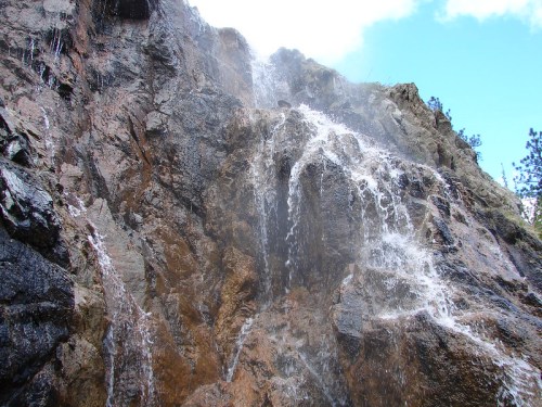 A cascading waterfall flows down a rocky cliff, surrounded by greenery and a blue sky.