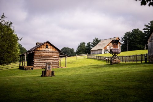 Two rustic log cabins on a grassy hillside, surrounded by trees and a wooden fence under a cloudy sky.