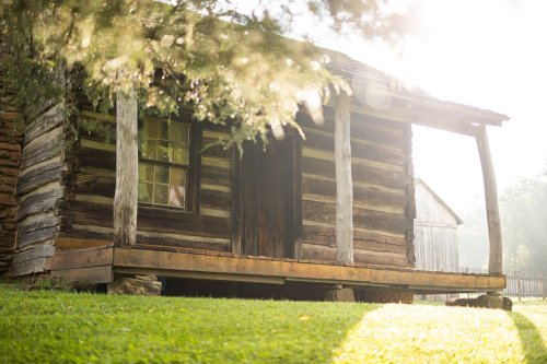 A rustic log cabin with a wooden porch, surrounded by green grass and trees, illuminated by soft sunlight.