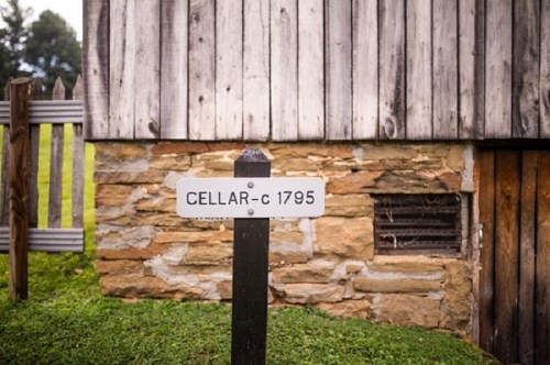 Sign reading "CELLAR-c 1795" in front of a wooden building with a stone foundation and grassy area.