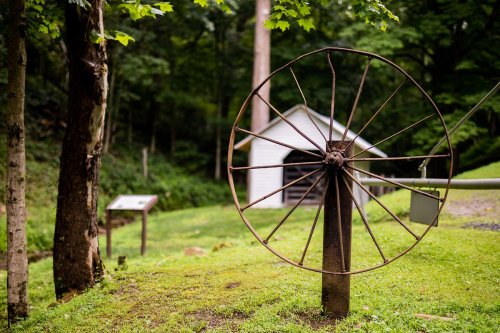 A rusty wheel on a post in a green landscape, with a white building and informational sign in the background.