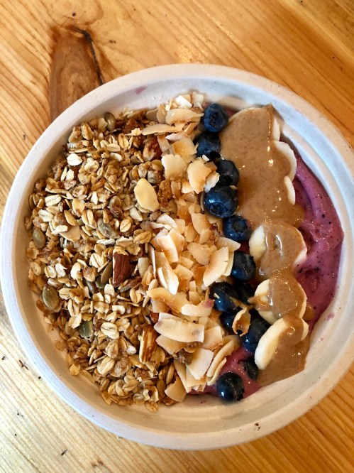 A bowl of smoothie topped with granola, sliced almonds, blueberries, and a drizzle of nut butter on a wooden table.
