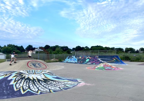 A colorful skatepark with vibrant murals and a skater on a ramp under a blue sky with scattered clouds.
