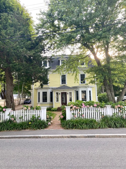 A charming yellow house with a white picket fence, surrounded by greenery and flowers, on a quiet street.