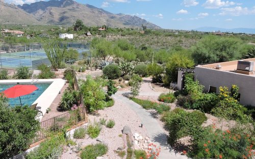 A scenic view of a garden with a pool, surrounded by mountains and tennis courts under a clear blue sky.