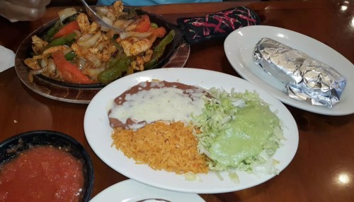 A plate of sizzling chicken fajitas with peppers, rice, refried beans, lettuce, and green sauce, alongside a wrapped burrito.