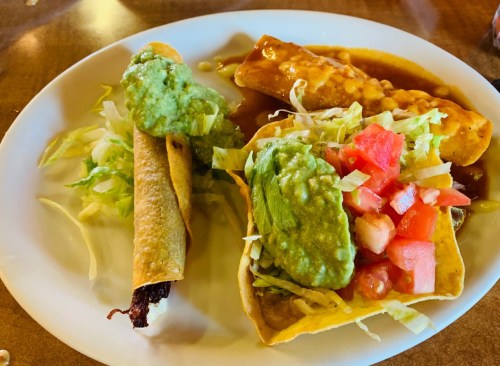 A plate of tacos and enchiladas topped with guacamole, lettuce, and diced tomatoes, served with a side of sauce.