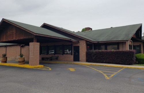 A single-story building with a sloped roof, brick exterior, and a small parking area in front.