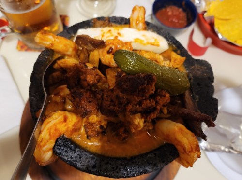 A sizzling stone bowl filled with shrimp, beef, cheese, and a green pepper, served with sides of salsa and tortilla chips.