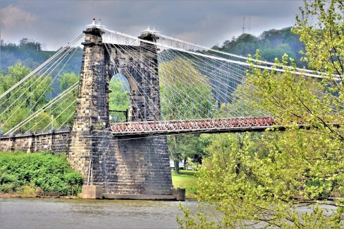 A suspension bridge with stone towers spans a river, surrounded by greenery and a cloudy sky.