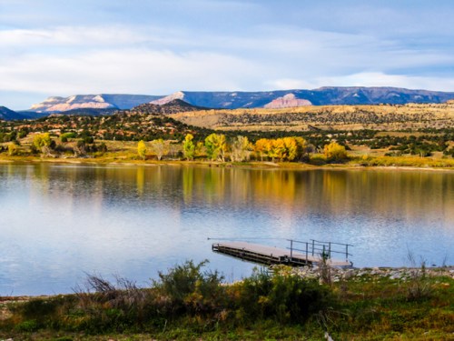 A serene lake reflects colorful trees and mountains under a cloudy sky, with a small dock in the foreground.