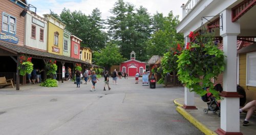 A lively outdoor scene in a theme park with colorful buildings, hanging plants, and visitors walking along the path.