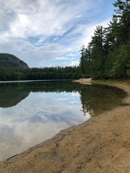 A serene lake surrounded by trees, with a sandy shore and a cloudy sky reflecting on the water's surface.