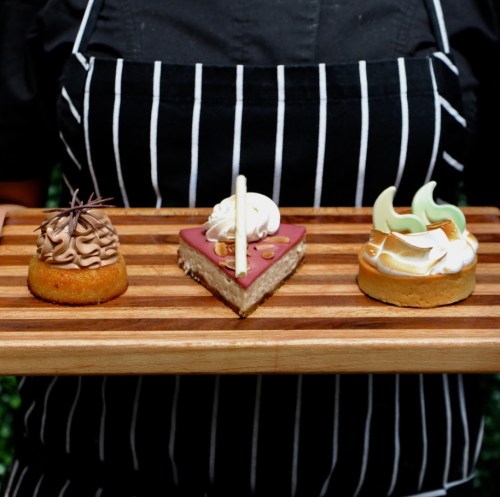 A person in a striped apron holds a wooden tray with three assorted desserts: a chocolate swirl, a berry tart, and a meringue treat.
