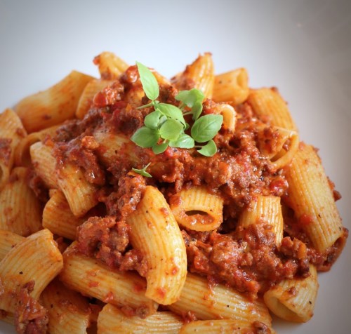 A plate of rigatoni pasta topped with a rich meat sauce and garnished with fresh basil.