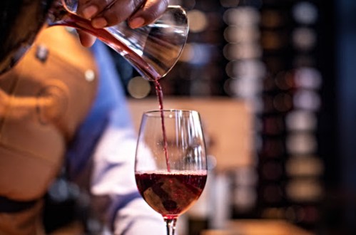 A hand pours red wine from a decanter into a glass, with a blurred background of wine bottles.